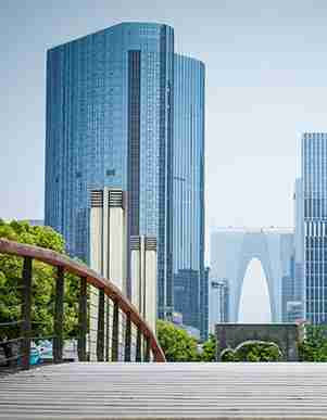 Modern cityscape with tall glass skyscrapers, a distinctive arch-shaped building in the background, and a wooden bridge with a railing in the foreground on a clear day.