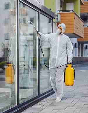 Person in protective gear spraying a building's glass exterior with a hose connected to a yellow container.