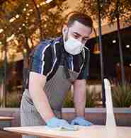 A person wearing a mask and gloves cleans a table in an outdoor setting.