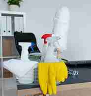 A basket of cleaning supplies, including spray bottles, yellow gloves, a duster, and a cloth, is on a table next to a keyboard. A shelf with binders is in the background.