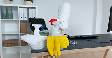Cleaning supplies, including spray bottles, a duster, and yellow gloves, are placed on a black desk in an office setting with shelves in the background.