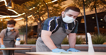 Two restaurant workers wearing masks and gloves clean tables.