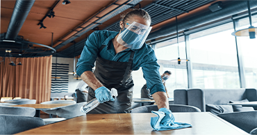 A person in a face shield and mask cleans a table in a restaurant with a spray bottle and cloth.