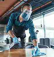 A person wearing protective gear cleans a table in a restaurant.