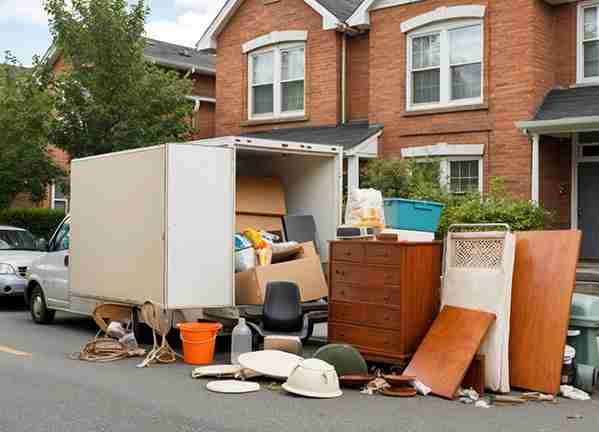A moving truck with its back open is parked on a residential street, with various household items and furniture placed on the ground nearby.