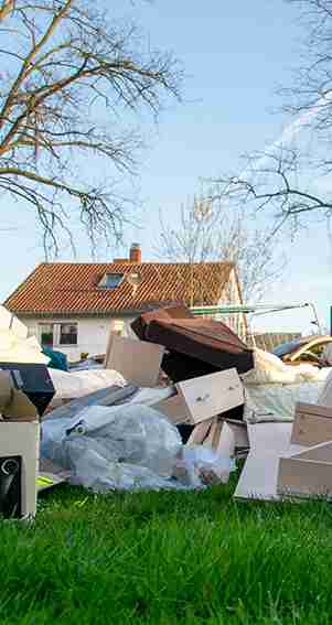 Piles of discarded cardboard boxes and plastic waste are scattered on a lawn in front of a house with a red-tiled roof.