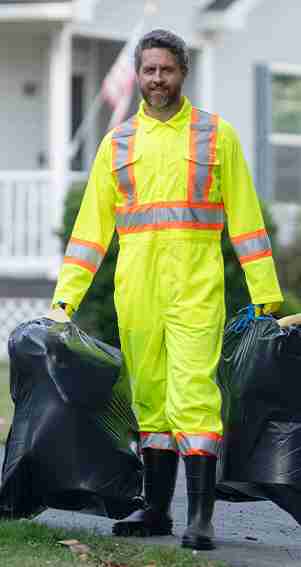 A person wearing a high-visibility yellow suit and black boots carries two large black garbage bags outside in a residential area.