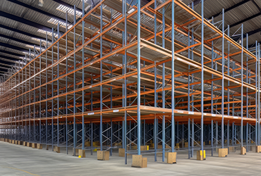 Large empty warehouse with multiple levels of orange and gray metal storage racks, concrete floor, high ceiling, and minimal lighting.