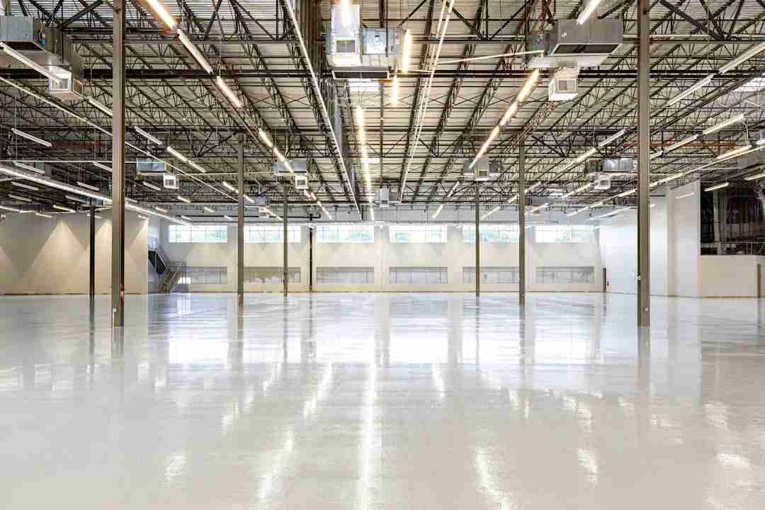 A large, empty industrial warehouse with polished white floors, high ceilings, exposed beams, and overhead lighting. Natural light enters through rectangular windows along the back wall.