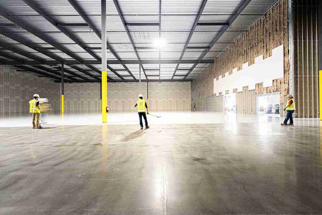 Three construction workers in safety vests and helmets stand inside a large, empty warehouse with unfinished walls, concrete floor, and exposed ceiling beams. The space is brightly lit by overhead lights.
