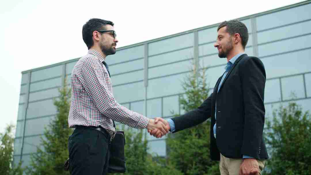 Two men in business attire are standing outside in front of a modern building, shaking hands. Trees and the building's glass façade are visible in the background.