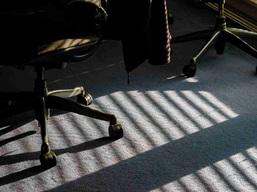Two office chairs on wheels sit on a carpeted floor. Sunlight streams through window blinds, casting striped shadows across the carpet and the chair bases.