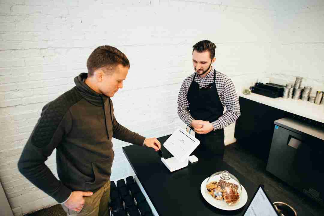 A man in a brown jacket uses a touchscreen payment terminal at a counter, while a man in a checkered shirt and apron stands behind the counter. A plate of pastries and a receipt holder are on the counter.