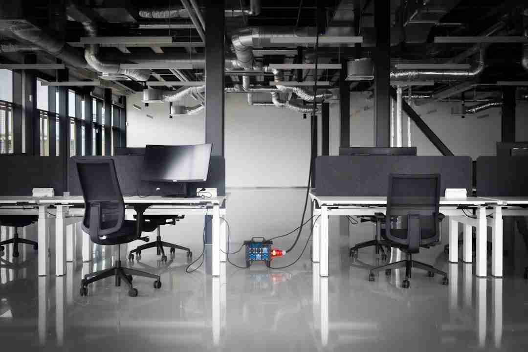 An empty modern office space with black chairs, white desks, computer monitors, and visible ceiling pipes. A power unit with cables is on the floor between the desks. The room has large windows on the left.