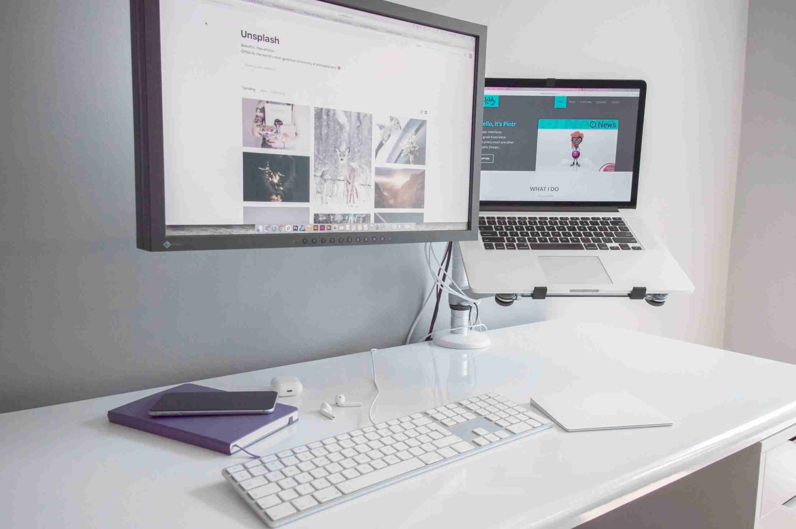 A minimalist white desk setup with a wireless keyboard, trackpad, notebook, and earphones. Two screens are present: a large monitor displaying a photo website and a laptop on a stand showing a web page.