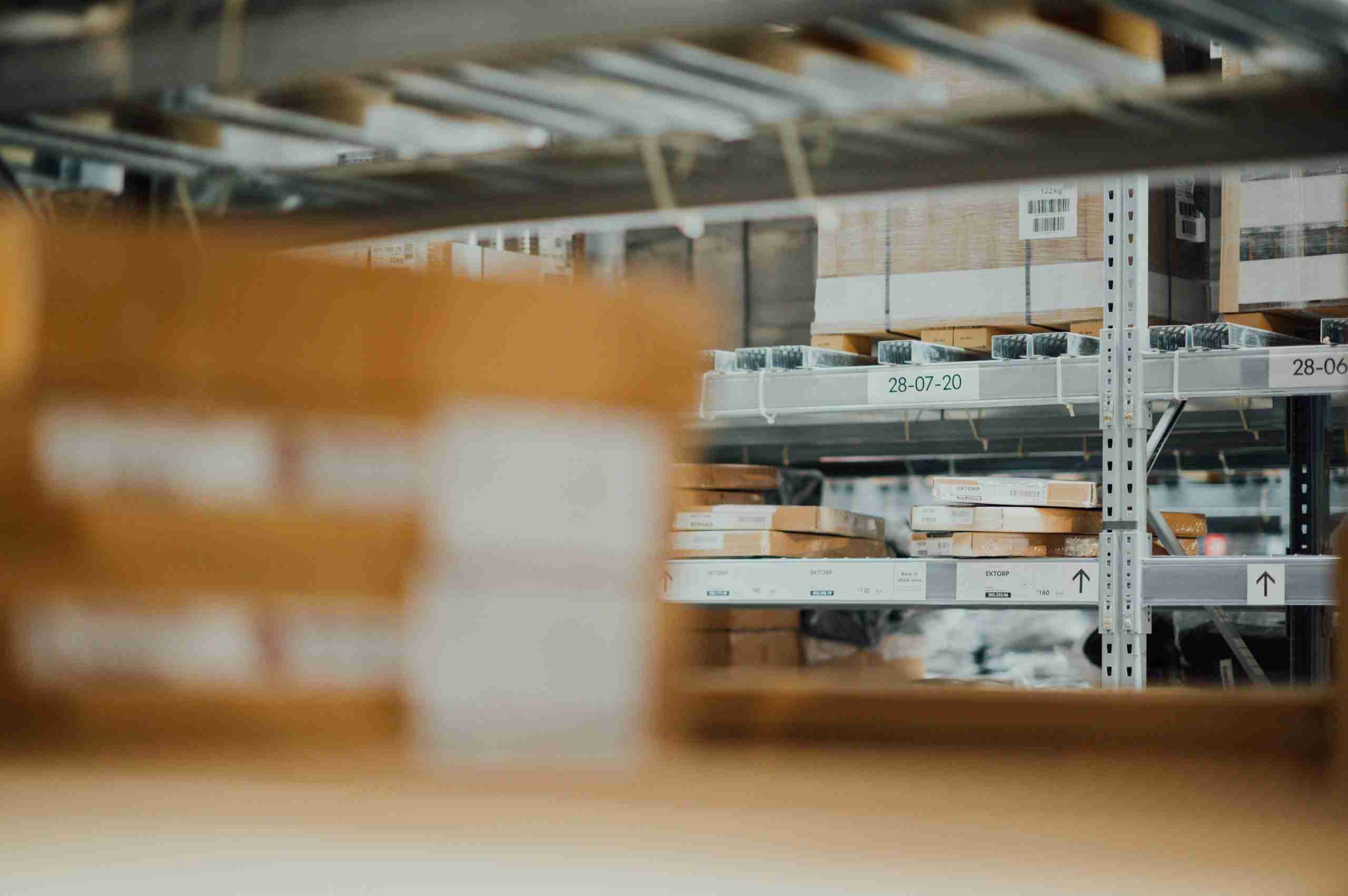 Shelves in a warehouse are filled with stacks of flat-packed cardboard boxes and other packages, organized and labeled with barcodes and dates. The foreground is blurred, focusing on the back shelves.