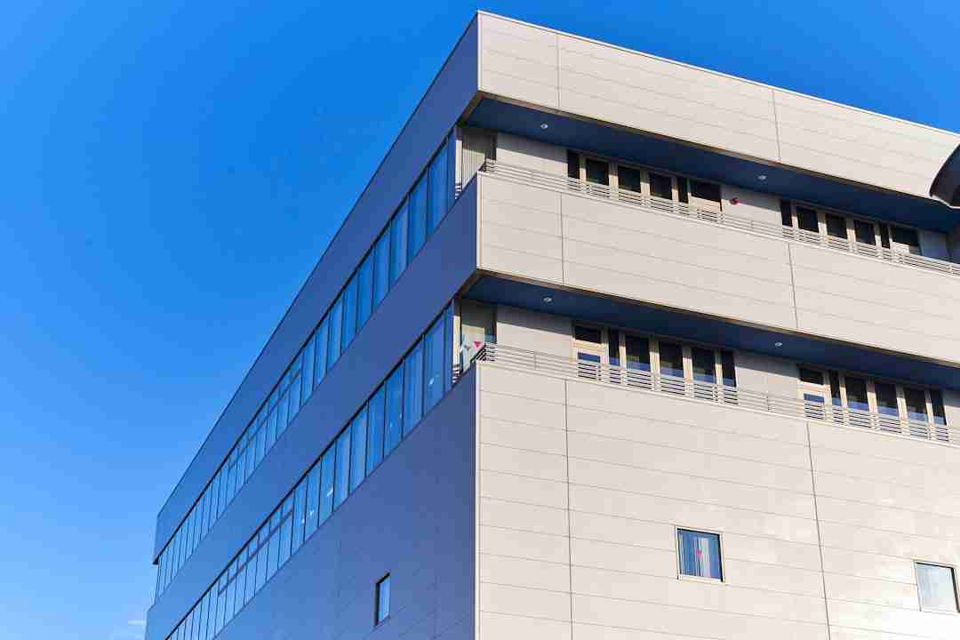 A modern, light gray building with large windows and a flat facade is pictured against a clear blue sky. The photo is taken from a low angle, showing two sides of the structure.