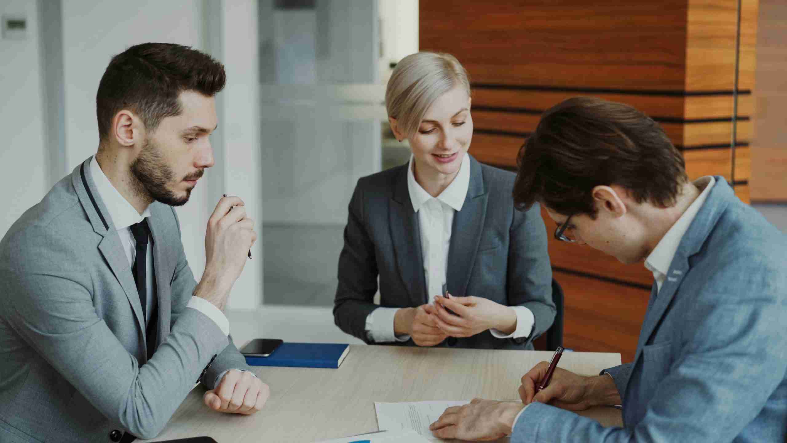 Three people in business attire sit at a table. One person is signing a document while the others observe. A phone and a blue folder are on the table. The setting appears to be a modern office.