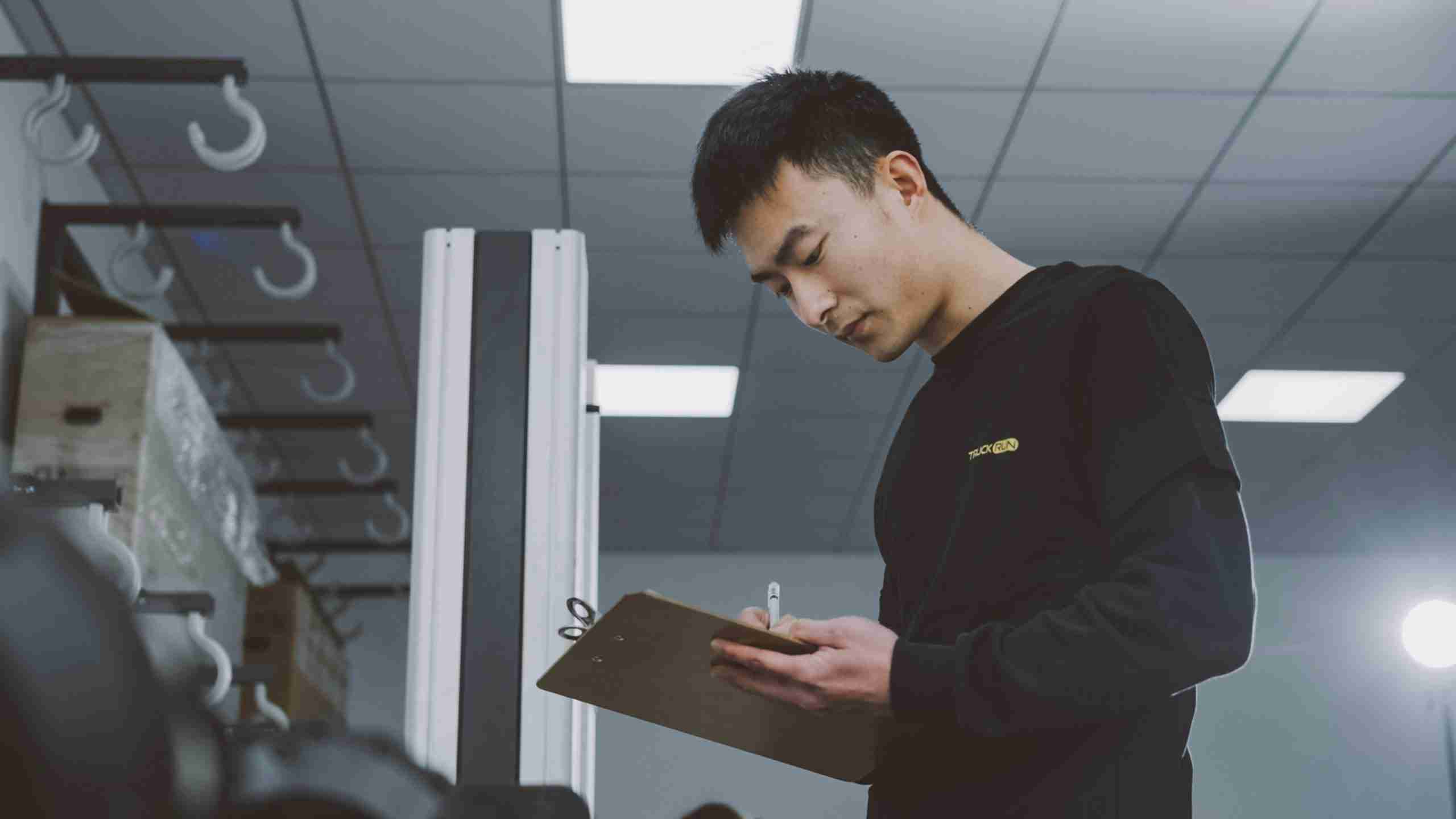 A person wearing a black shirt stands indoors, looking down and writing on a clipboard. The background shows part of an office or lab with ceiling lights and equipment.