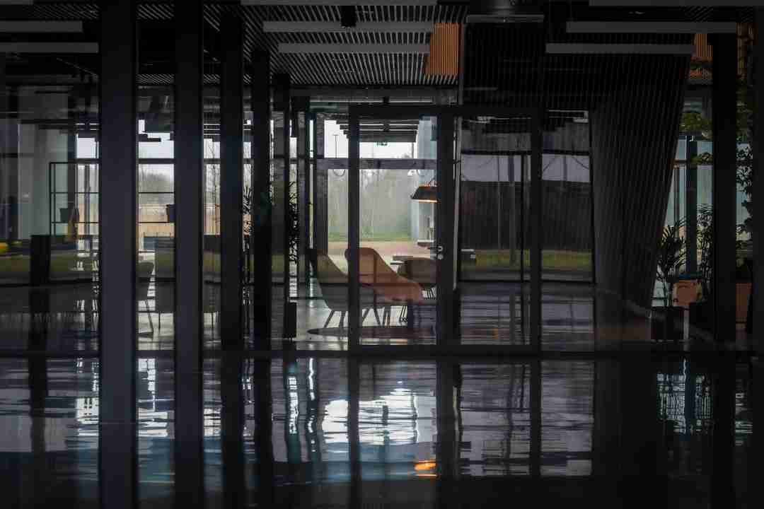 Interior view through glass walls shows empty modern office space with reflections on a shiny floor, a lounge chair, a small table, and large windows revealing an outdoor scene with greenery in the background.
