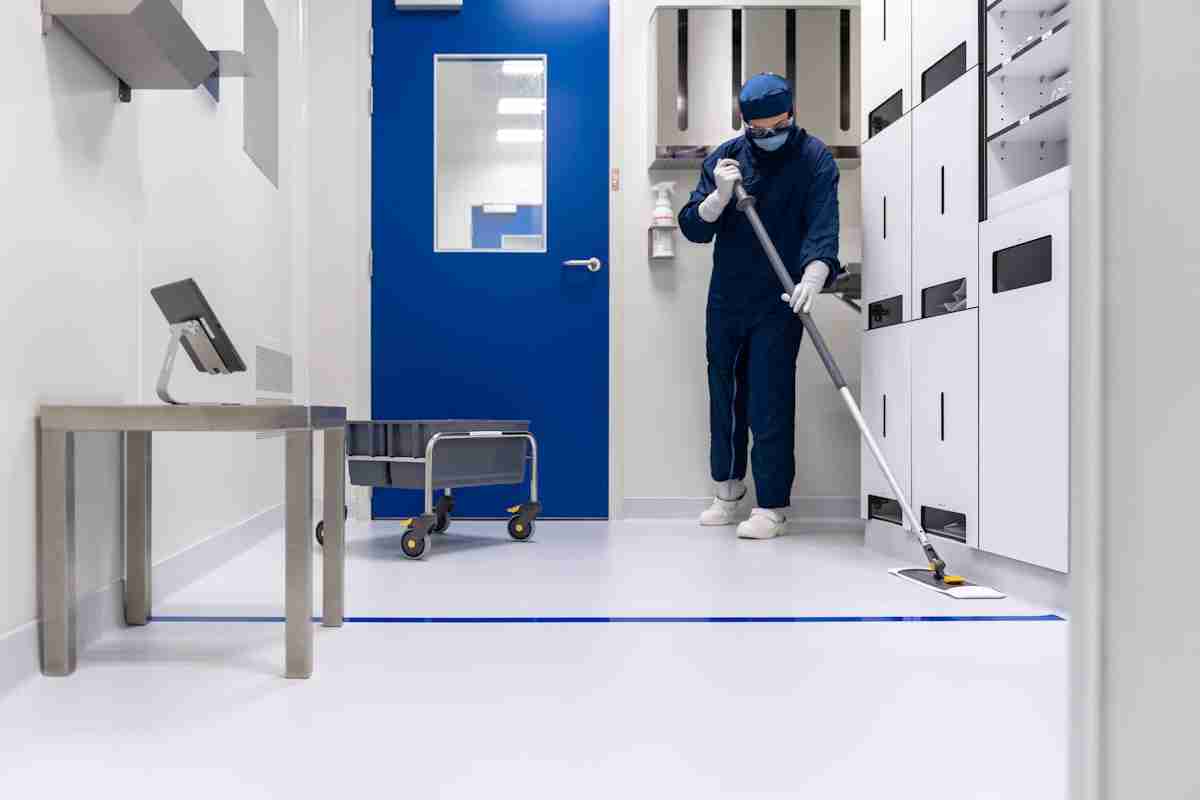 A person in protective clothing and a face mask uses a mop to clean the white floor of a sterile room with white cabinets, a blue door, and a metal cart nearby.