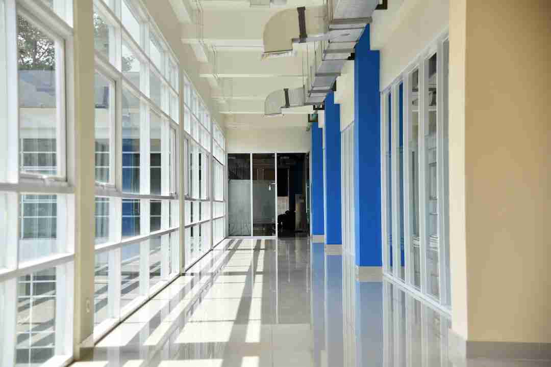 A long, empty hallway with large floor-to-ceiling windows on one side, reflecting sunlight onto the shiny tile floor. Walls are cream and blue, and exposed ductwork runs along the ceiling. Glass doors are at the far end.