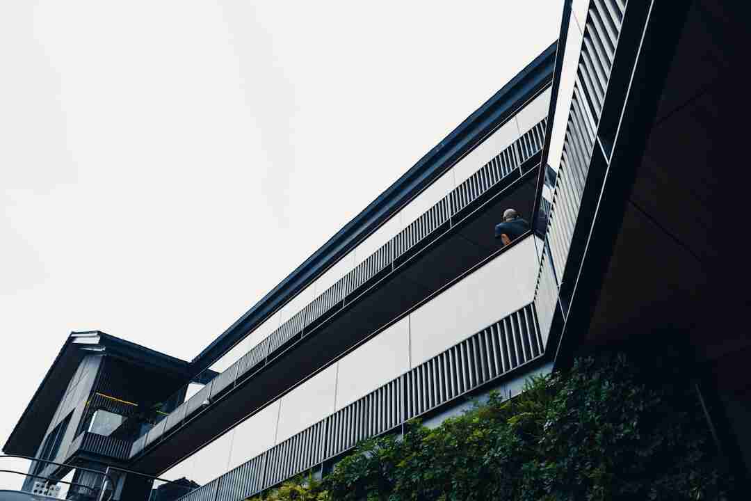 A person sits on the upper balcony of a modern multi-story building with metal railings and large windows. Green plants are visible on the lower level, and the sky is overcast.