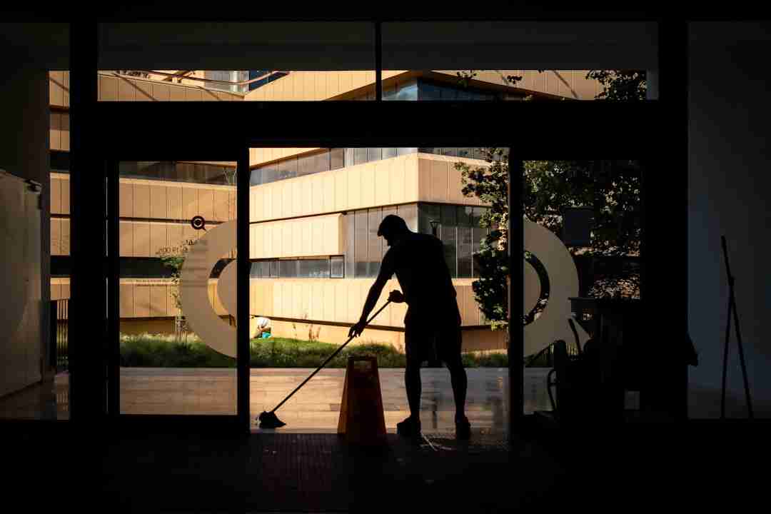 A person is mopping the floor near the entrance of a building with a caution wet floor sign placed nearby. Sunlight and reflections from modern buildings can be seen outside through the glass doors.