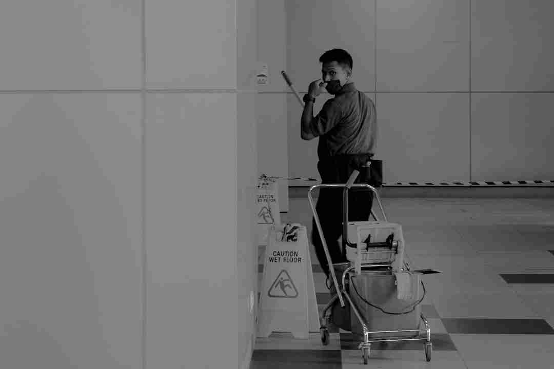 A janitor wearing a mask stands next to a cleaning cart and wet floor signs in a hallway, holding a tool and looking toward the camera. The scene is lit by overhead lights and has tiled flooring.