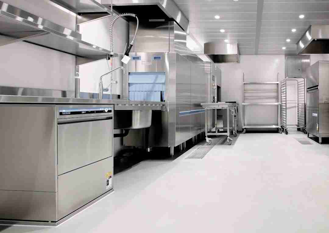 A modern commercial kitchen with stainless steel appliances, countertops, and shelves. There is a dishwasher, industrial sink, preparation area, and empty racks against the wall. The space is clean and brightly lit.