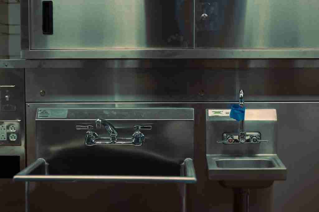 A stainless steel commercial sink setup with a large deep sink on the left and a smaller handwashing sink with a blue label and push faucet on the right. The background shows stainless steel cabinets and equipment.