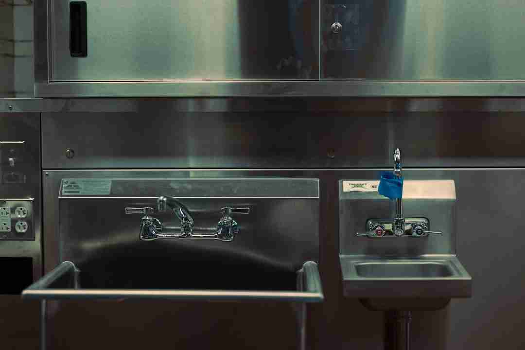 A stainless steel commercial sink setup with a large deep sink on the left and a smaller handwashing sink with a blue label and push faucet on the right. The background shows stainless steel cabinets and equipment.