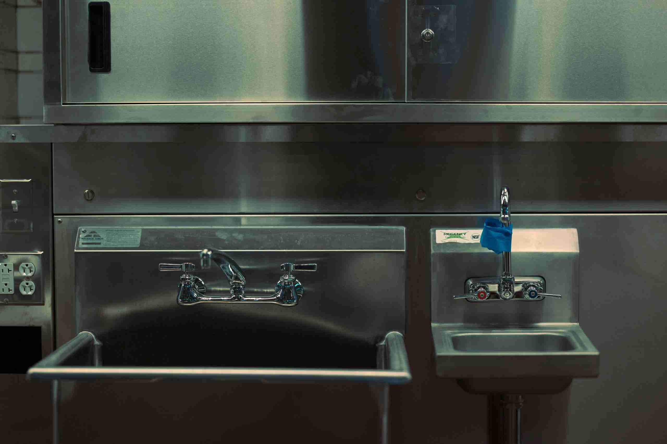 A large stainless steel sink with a double-handle faucet is next to a smaller handwashing sink with a single-handle faucet wrapped in blue tape. Both are set against a metallic wall in a commercial kitchen.