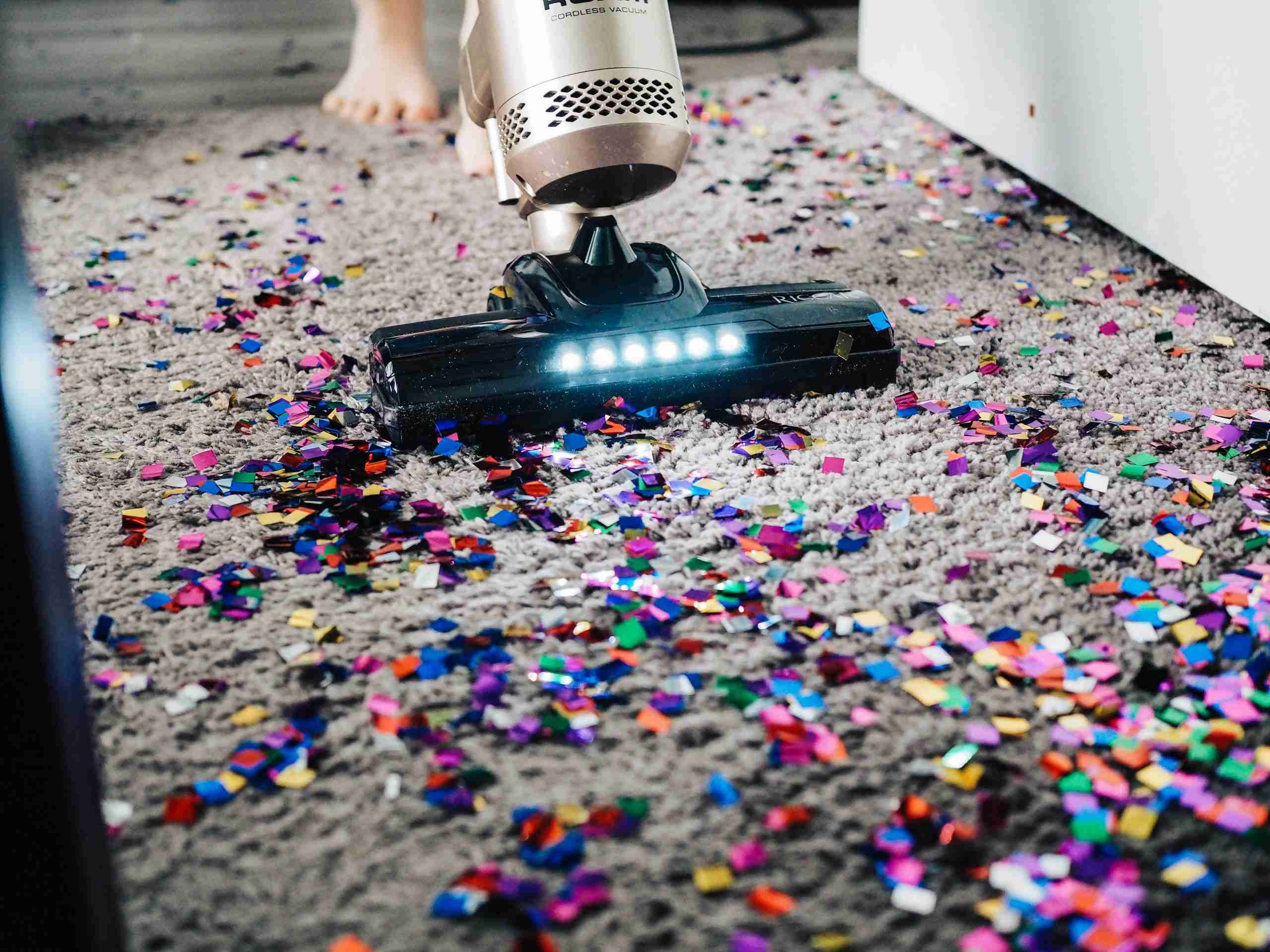 A person uses a vacuum cleaner with LED lights to clean up colorful confetti scattered on a gray carpet. Part of a white piece of furniture and a bare foot are visible in the background.