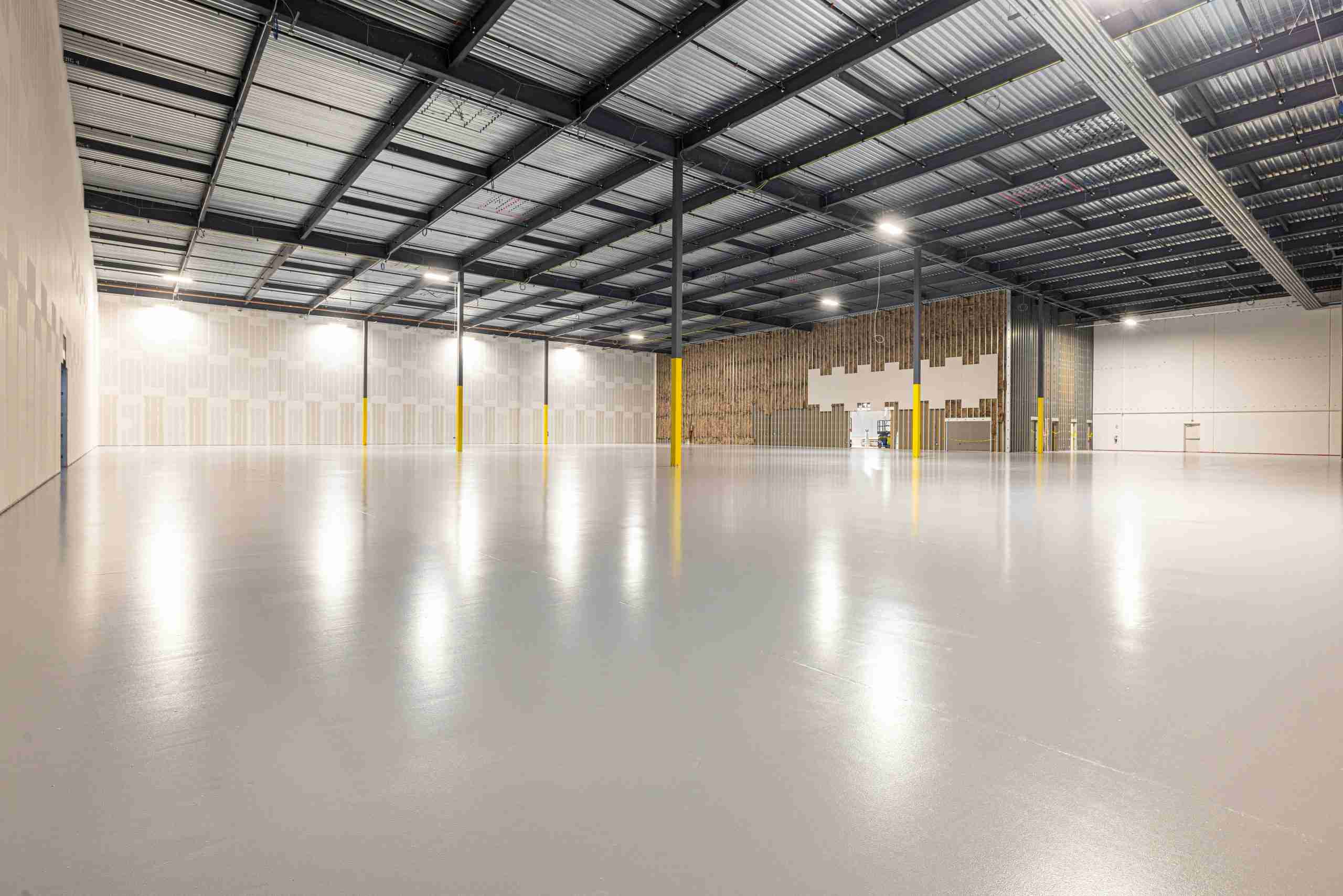 A large, empty warehouse with polished concrete floors, high ceilings, exposed beams, and yellow safety bollards around support columns. The space is brightly lit and has partially finished interior walls.