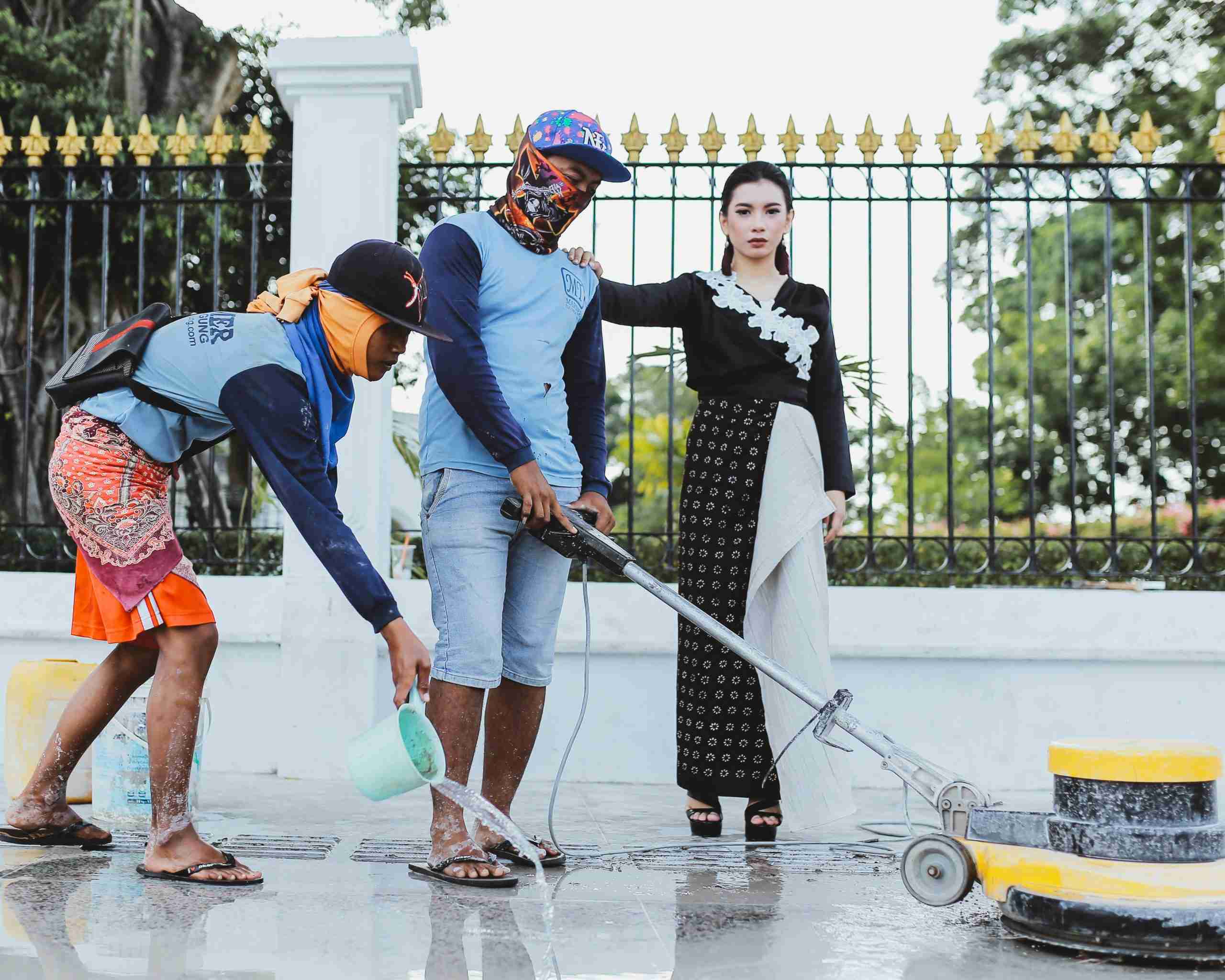 Two workers clean a sidewalk with water and a floor polishing machine while a woman in formal attire stands nearby, looking at the camera, in front of a decorative metal fence.
