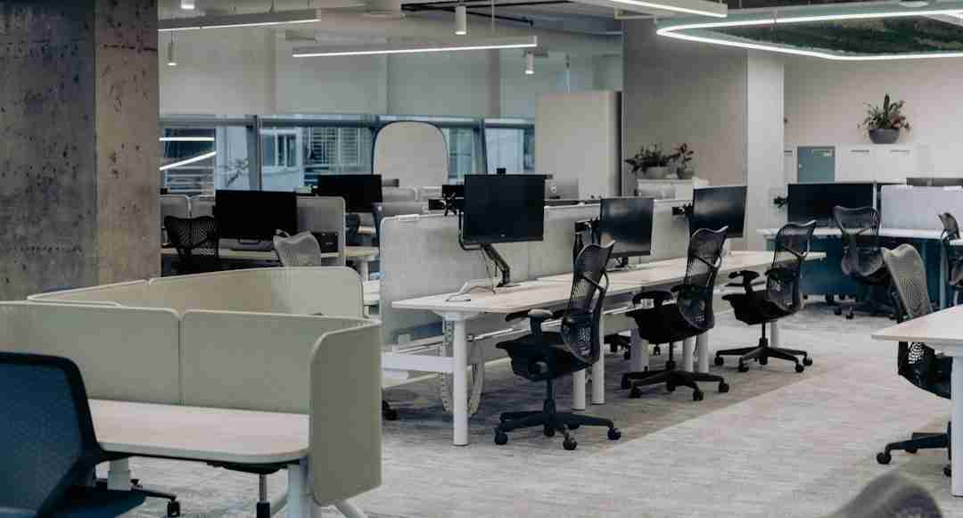Modern open-plan office with several empty workstations, ergonomic chairs, computer monitors, and partition panels. Large windows provide natural light. The space appears tidy and quiet.