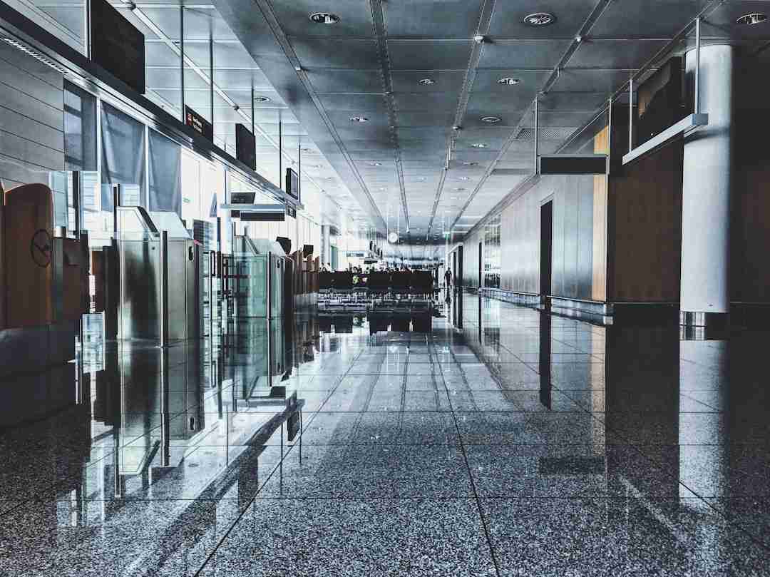 A modern airport terminal with shiny, reflective floors, empty check-in counters on the left, seating areas in the distance, and glass walls letting in natural light. The space appears clean and quiet.