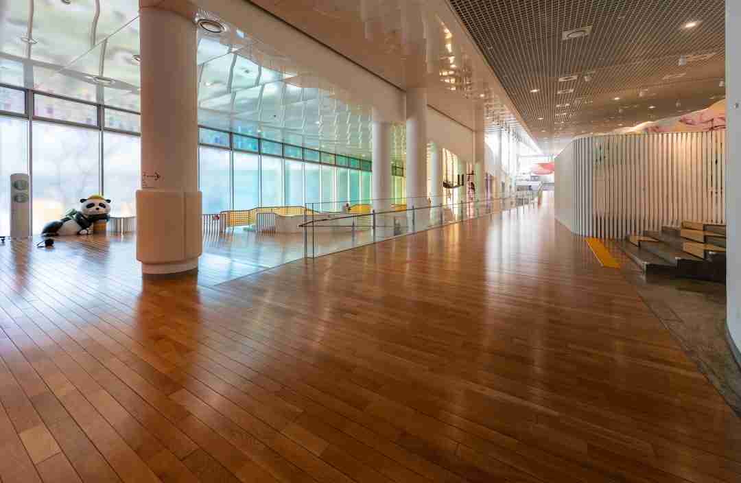 A spacious hallway with shiny wooden floors, large glass windows on the left, and white columns. A panda statue sits near the window. On the right, white vertical slats and steps are visible. The area is well-lit and empty.