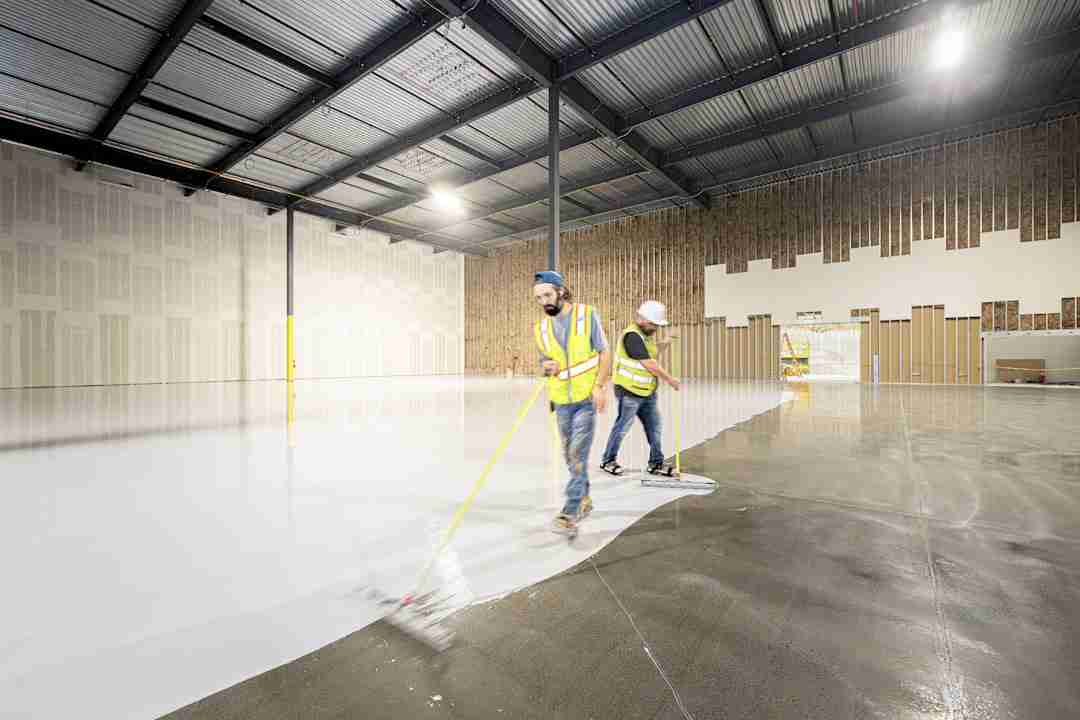 Two construction workers wearing safety vests and helmets apply a white coating to a large concrete floor inside a spacious industrial building with unfinished walls and a metal ceiling.