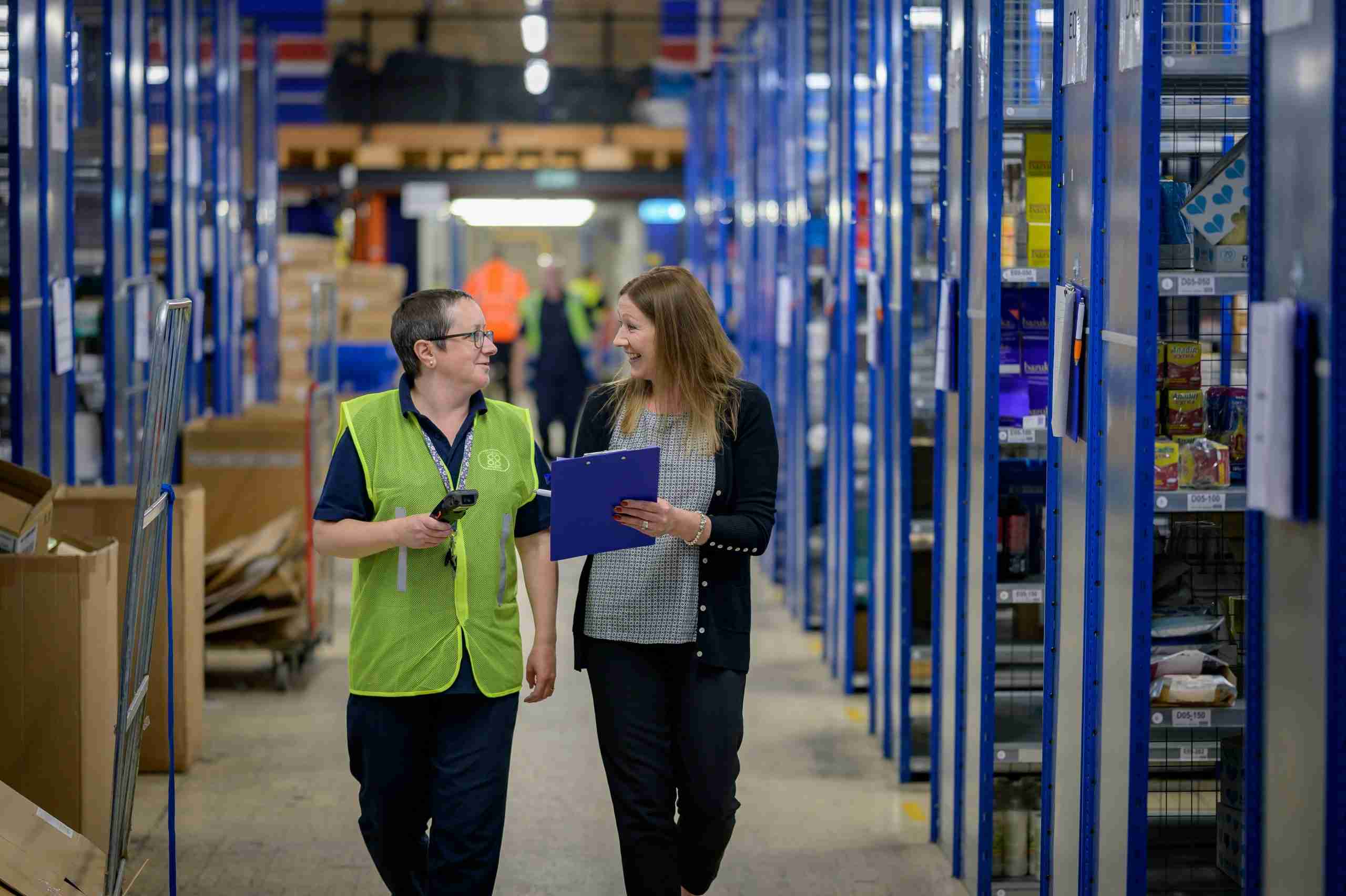 Two people walk down an aisle in a warehouse, one wearing a green safety vest and holding a handheld scanner, the other holding a blue clipboard. They appear to be having a conversation surrounded by shelves and boxes.