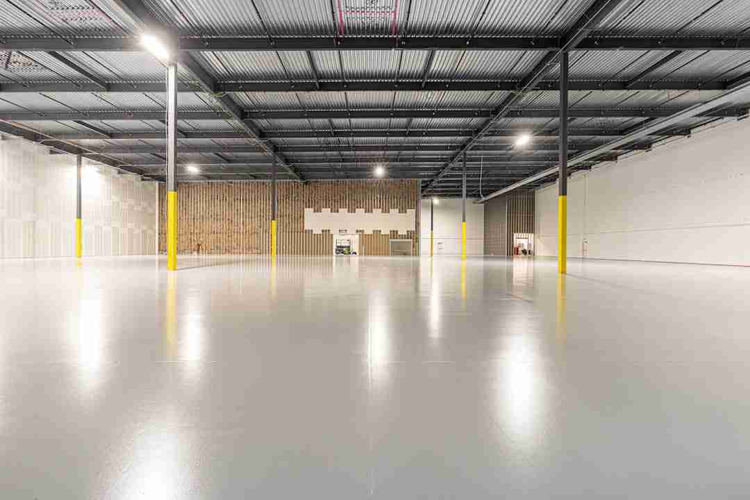 A large, empty industrial warehouse with polished concrete floors, high metal ceilings, yellow support columns, and unfinished walls. A small lift vehicle is parked near the back wall under bright overhead lighting.