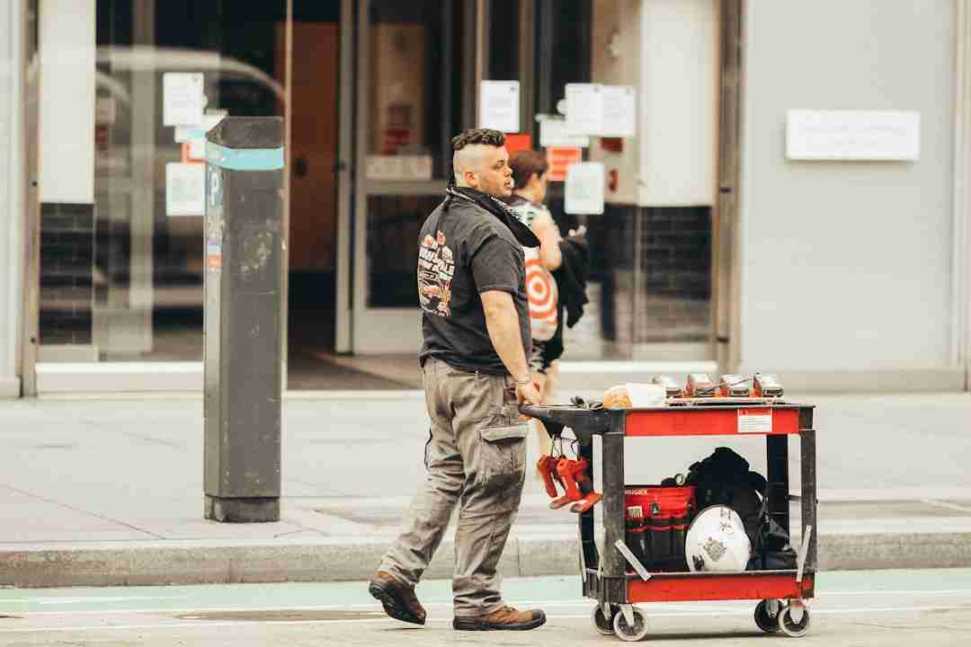A man in work clothes pulls a red and black utility cart loaded with tools and a helmet along a city street, with a building entrance and another person in the background.