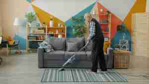 An older man vacuuming a striped rug in a modern living room with colorful geometric wall art, a gray sofa, and shelves with books and plants.