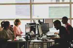 Four people are working at desks with computers in a modern office space with large windows. They appear focused and engaged with their screens and paperwork. The lighting is bright, and the atmosphere looks collaborative.