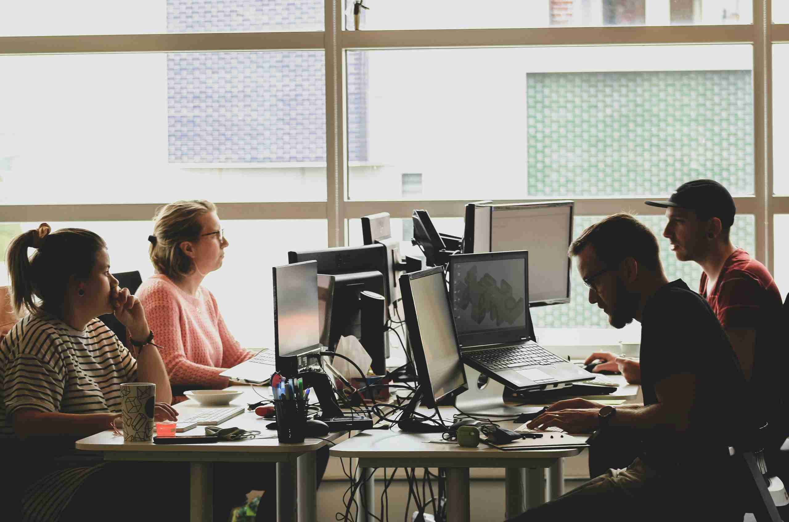 Four people are working at desks with computers in a modern office space with large windows. They appear focused and engaged with their screens and paperwork. The lighting is bright, and the atmosphere looks collaborative.