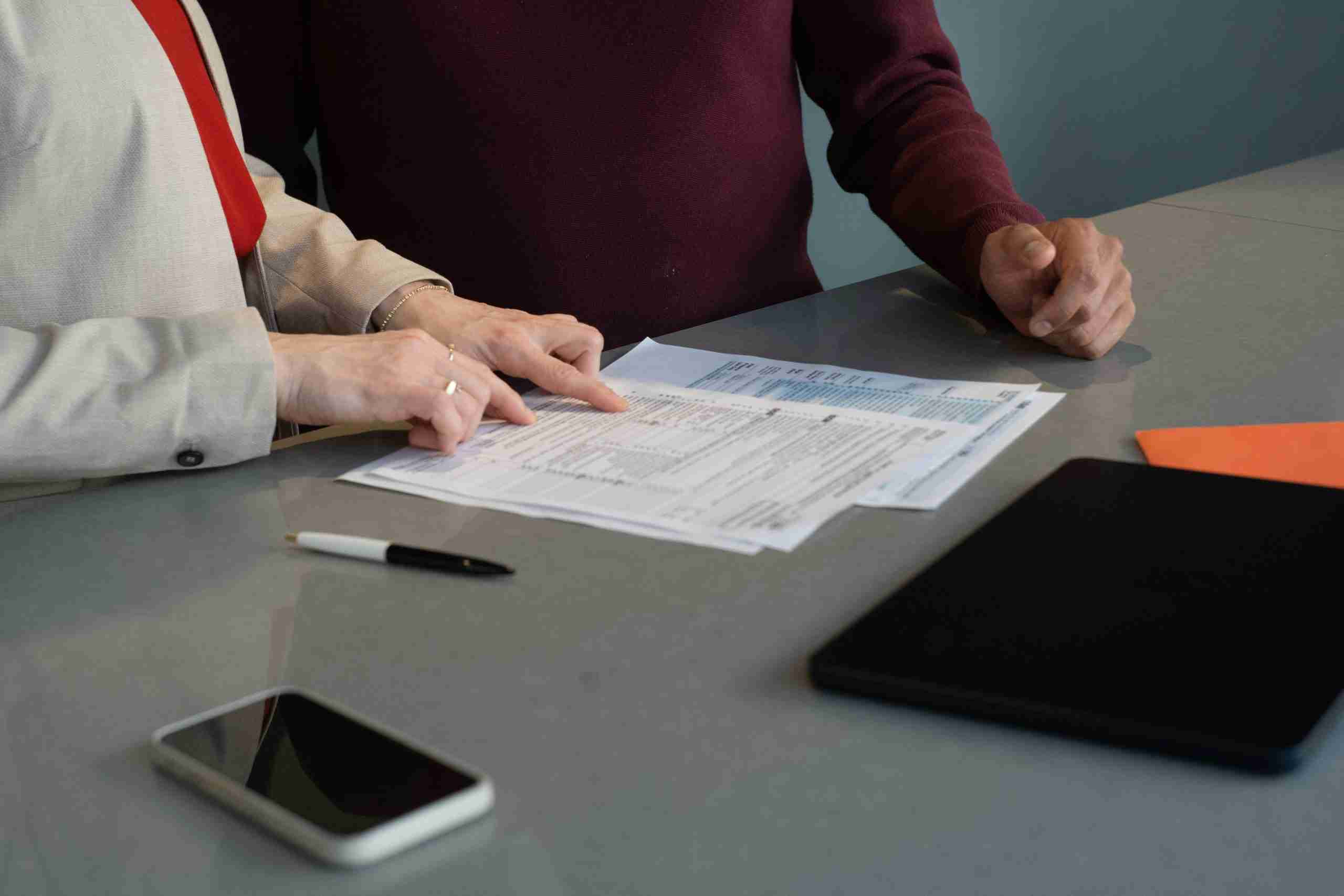 Two people review documents on a table, with one person pointing at the papers. A pen, smartphone, closed laptop, and orange envelope are also on the table. The focus is on their hands and the documents.