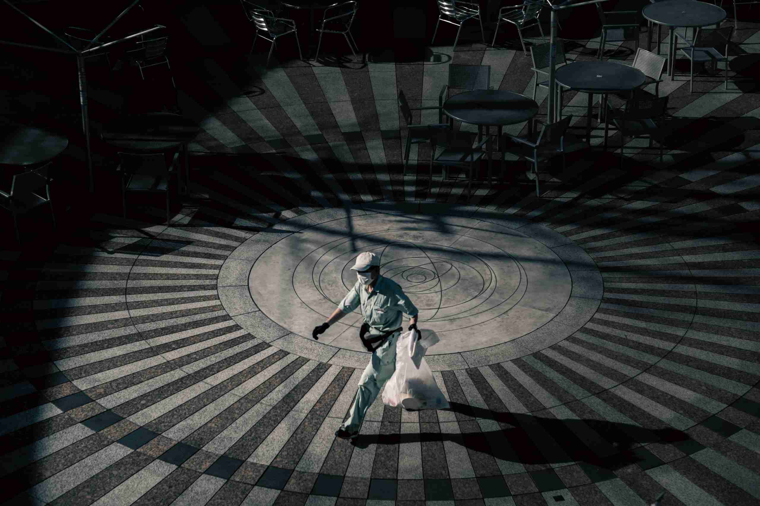 A person in uniform carries a white bag while walking across a circular, patterned plaza with metal tables and chairs arranged around the edge.