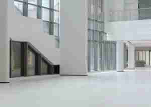 A spacious, empty interior of a modern building with large glass windows, white walls, and a polished white floor. Stairs are visible on the left, and a glass balcony lines the upper level.