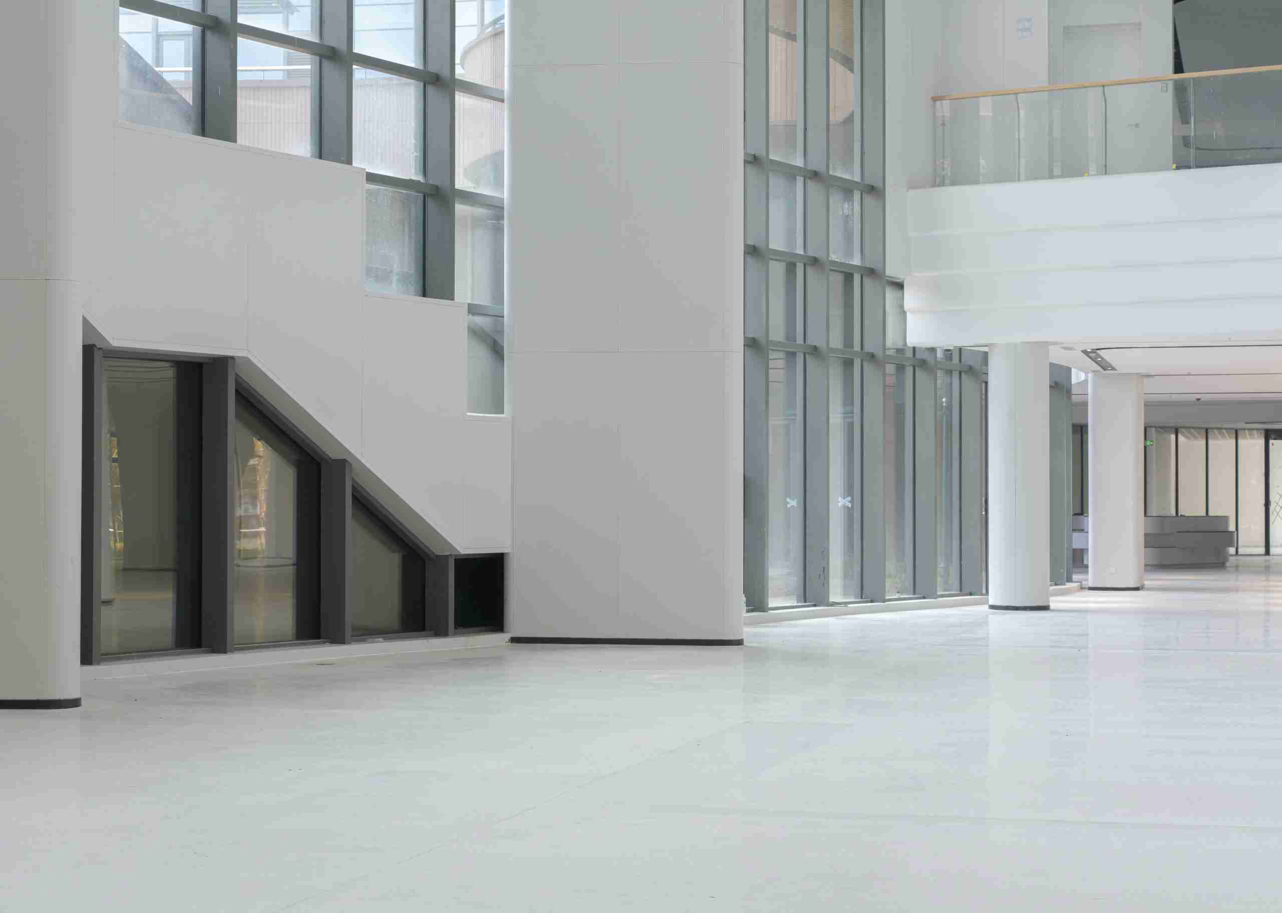 A spacious, empty interior of a modern building with large glass windows, white walls, and a polished white floor. Stairs are visible on the left, and a glass balcony lines the upper level.